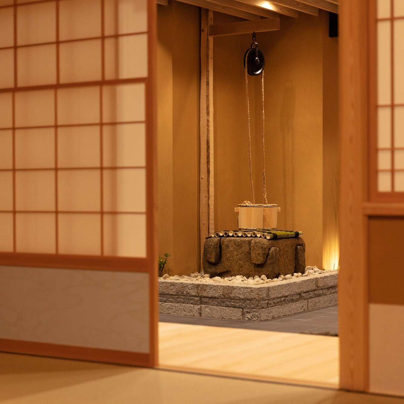 Seen through a paper-screen opening, a stone well with a rope pulley and wooden buckets stands against an earthen wall.
