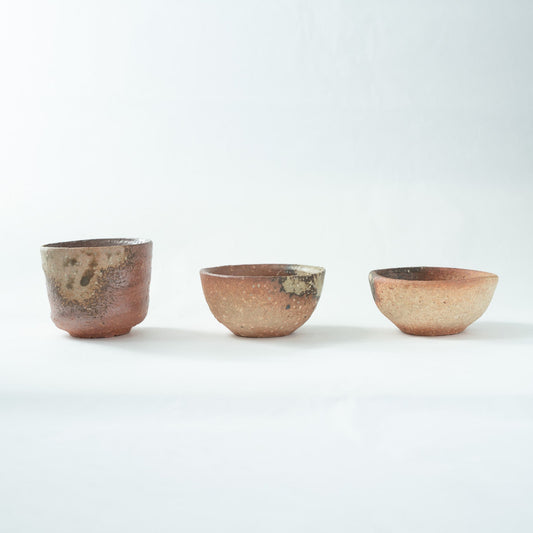 A slightly turned front view of the three Shigaraki sake cups, highlighting the scorch marks on the left cup and the textured surfaces of the other two bowls.