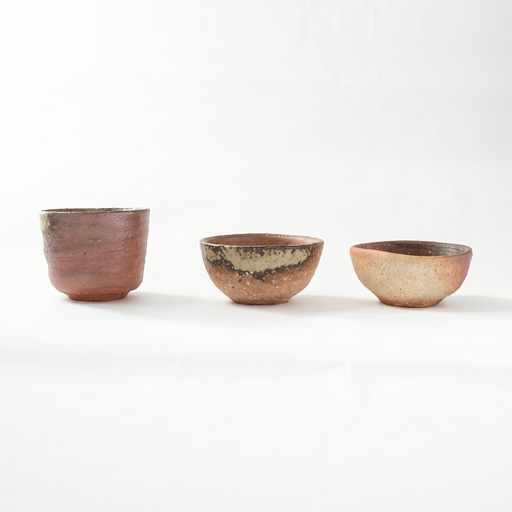 Front view of three distinct Shigaraki ware GUINOMI by Rakusai Takahashi arranged in a row, showing a tall cylindrical cup, a medium bowl with green ash glaze, and a smaller earthy red cup.