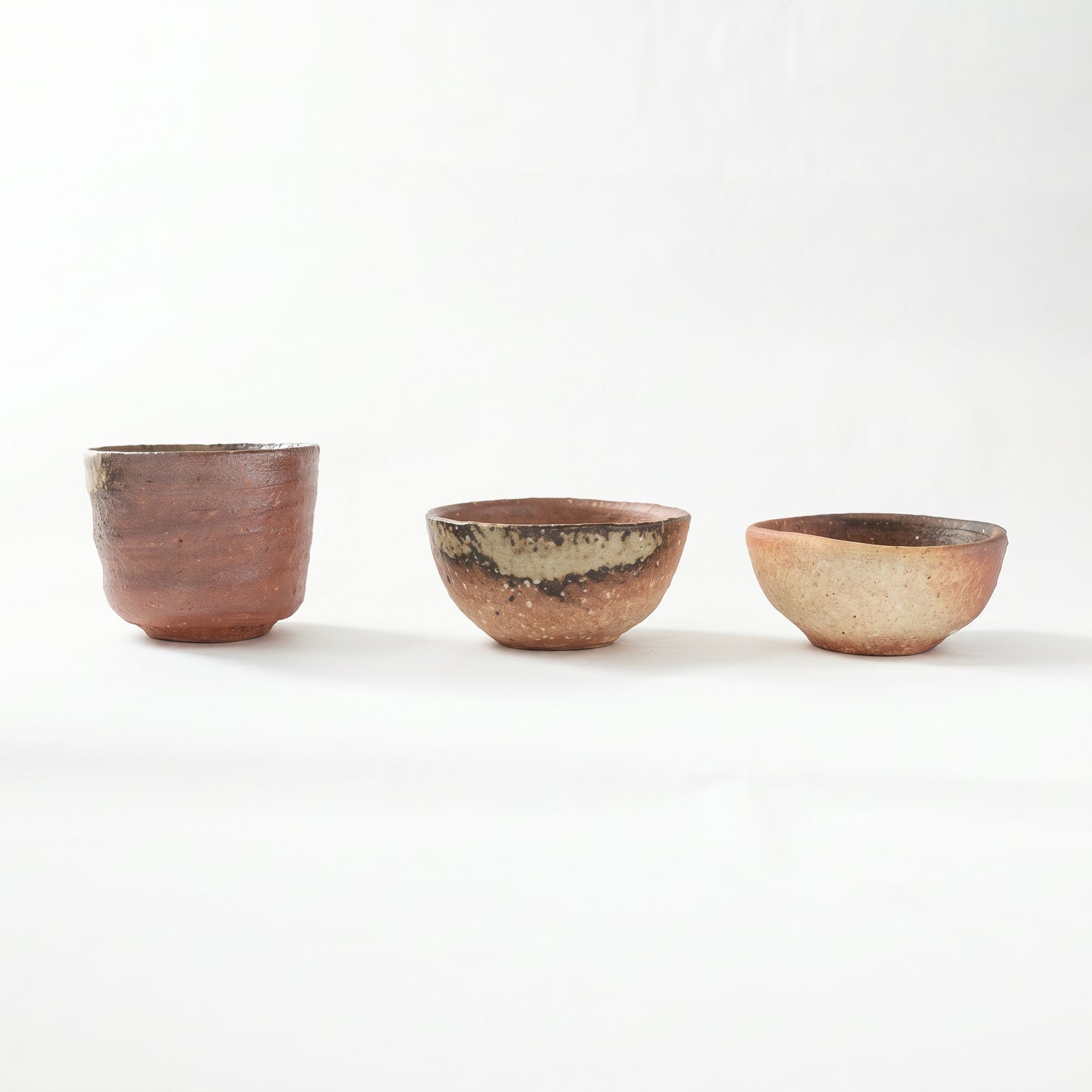 Front view of three distinct Shigaraki ware GUINOMI by Rakusai Takahashi arranged in a row, showing a tall cylindrical cup, a medium bowl with green ash glaze, and a smaller earthy red cup.