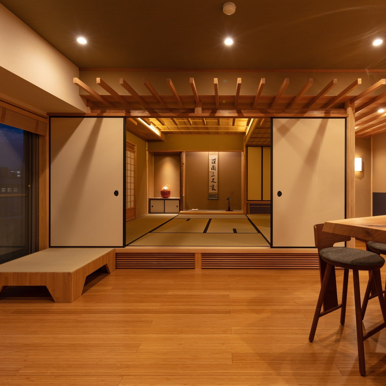 Raised tatami room glowing at dusk beyond an open-plan salon, showing the layered spaces of contemporary Japanese architecture.