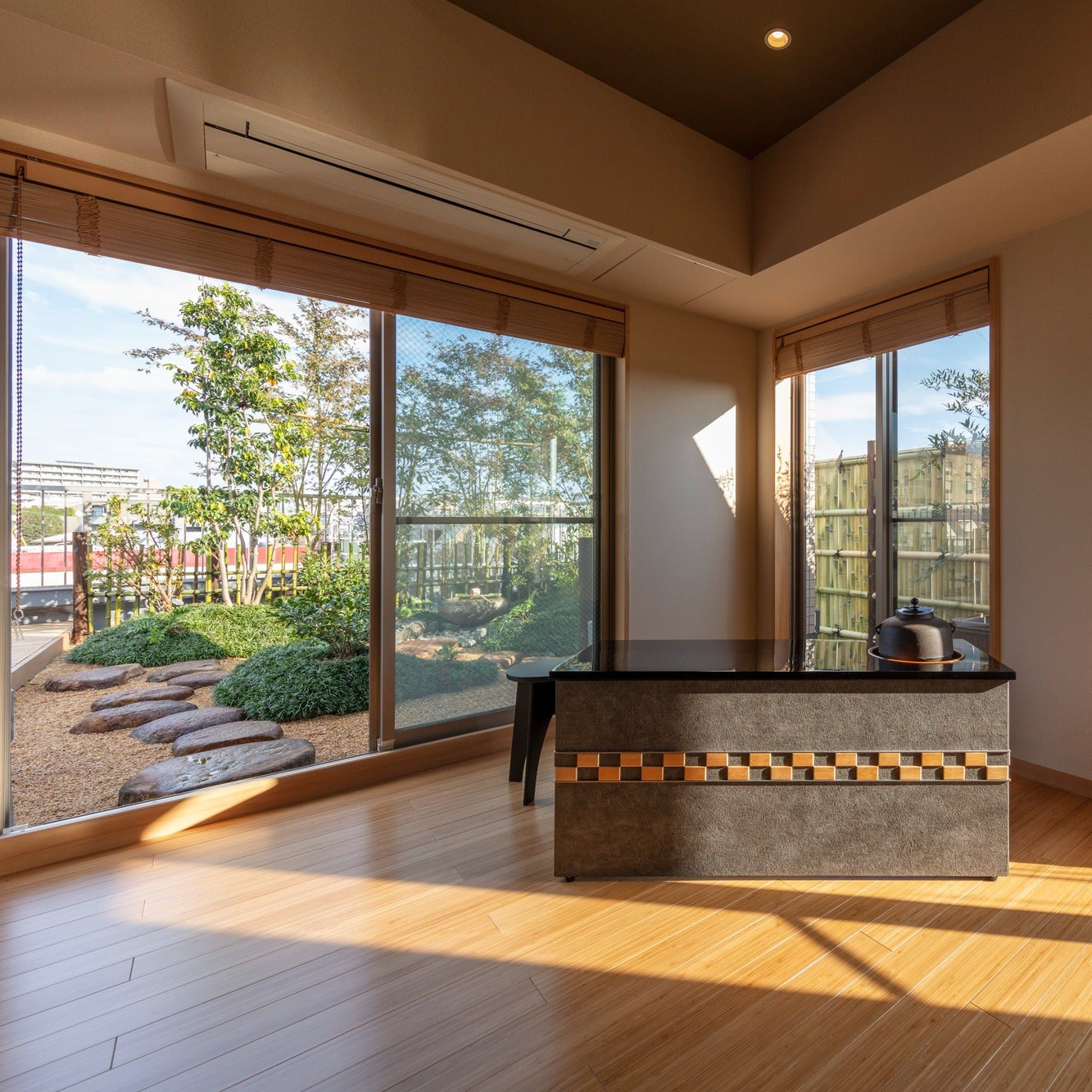 A low hearth island with an iron kettle before wide windows opening to a rooftop garden of maple and stepping stones.