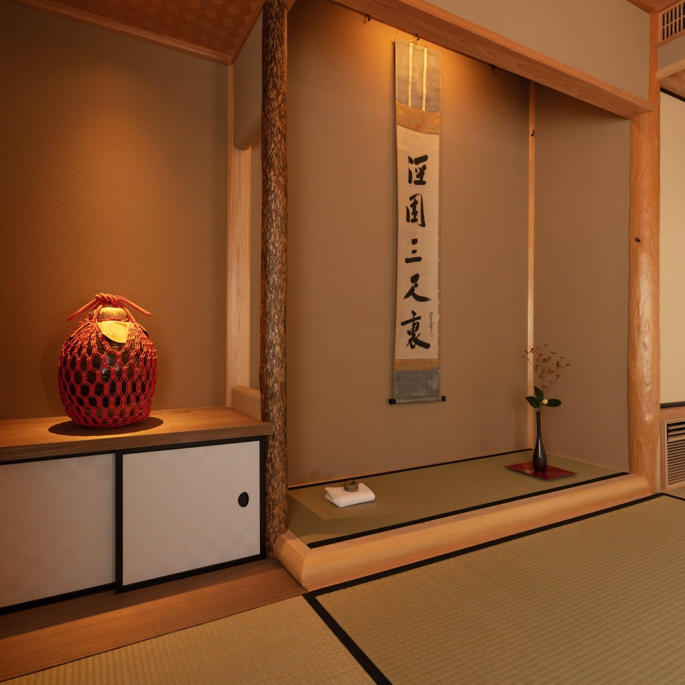 Tokonoma alcove with a rope-netted gold vessel, a hanging calligraphy scroll and a single flower beside a natural log post.