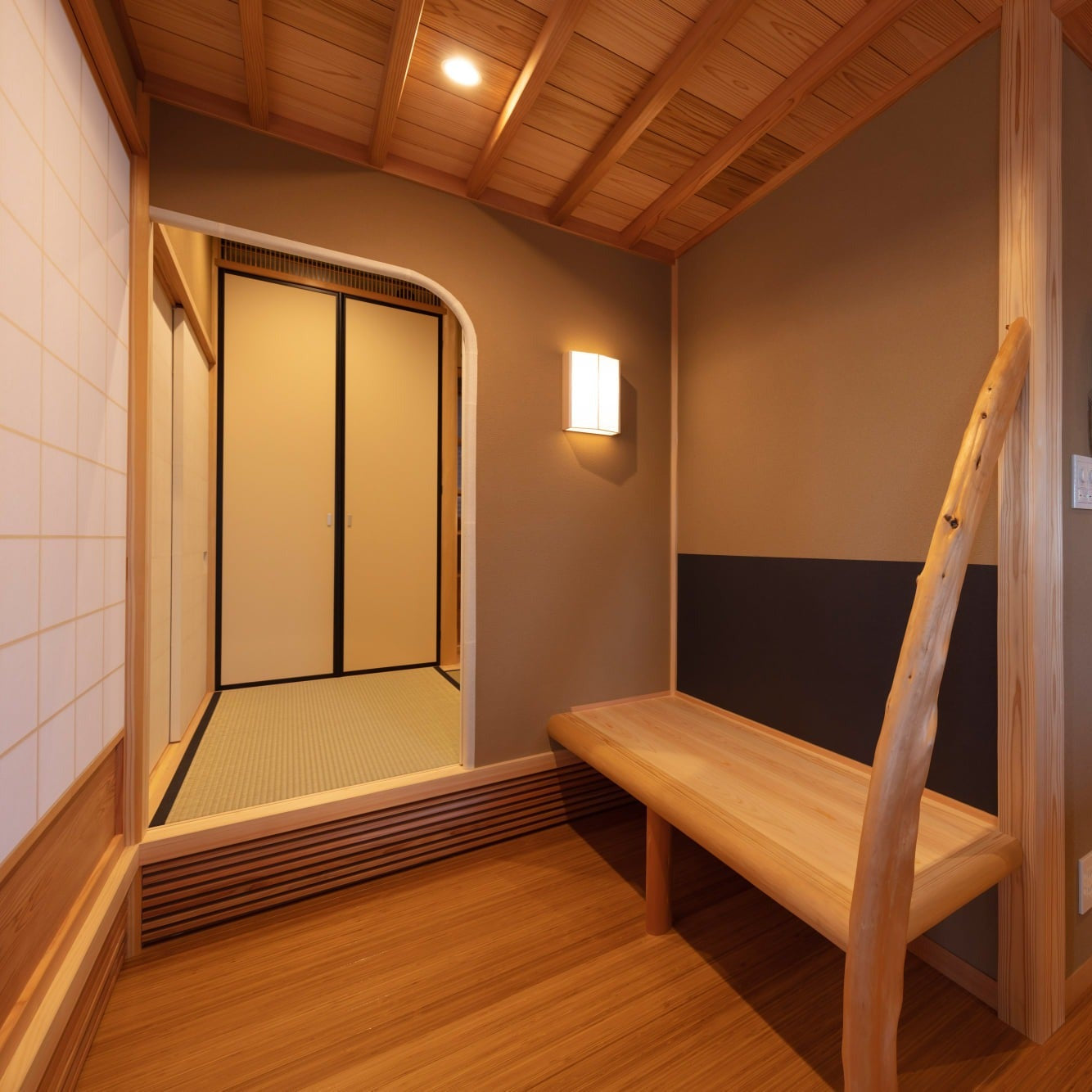 Waiting area with a cedar bench, a natural-edge post and an andon wall light beside an arched threshold to the tatami room.