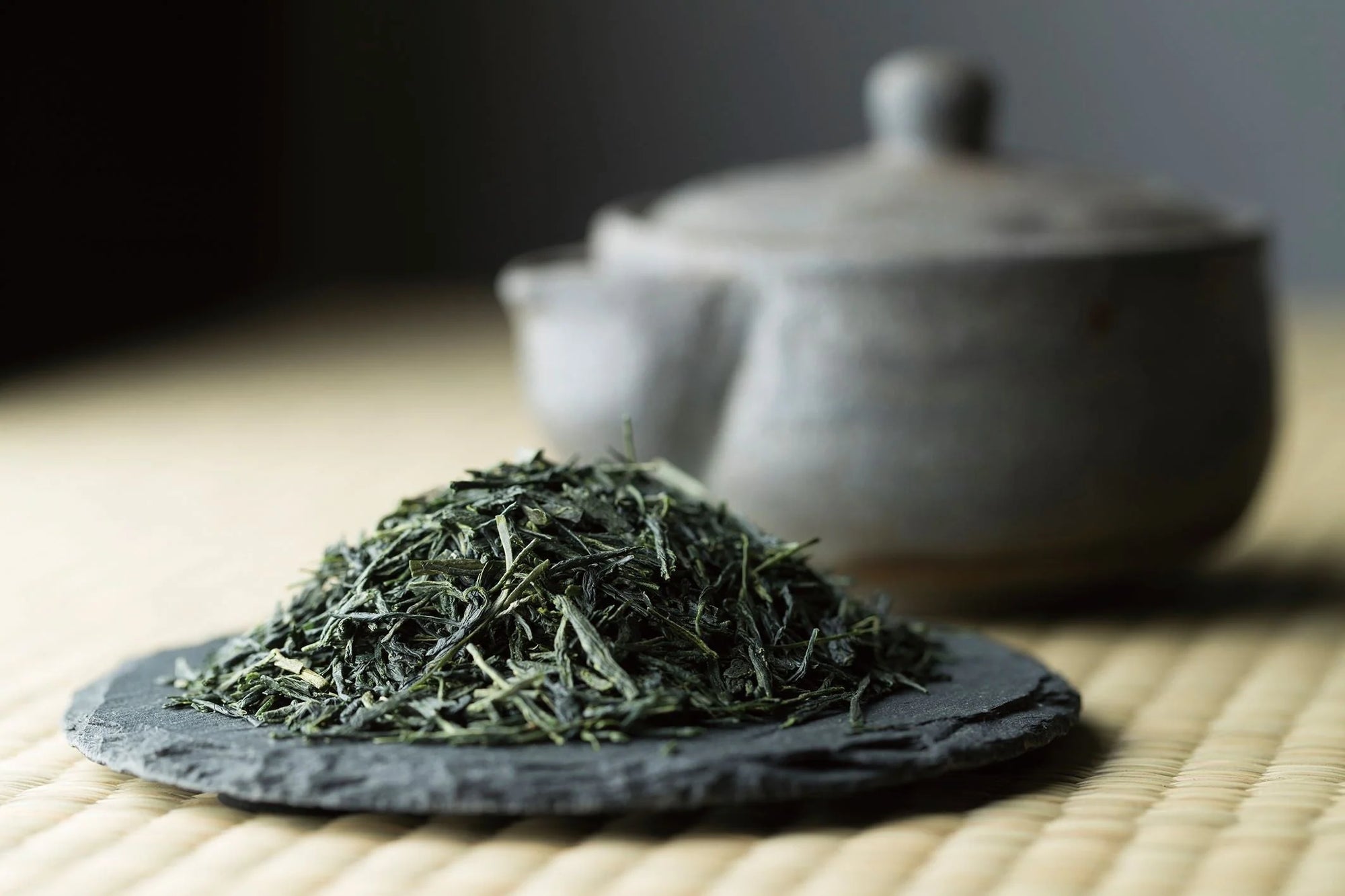 A mound of premium dry green tea leaves on a dark slate plate with a traditional Japanese clay teapot softly blurred in the background.
