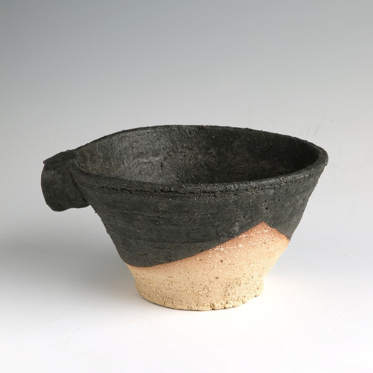 Front view of the wide black spouted vessel on a grey ground, the granular black upper body above a band of bare sandy clay.