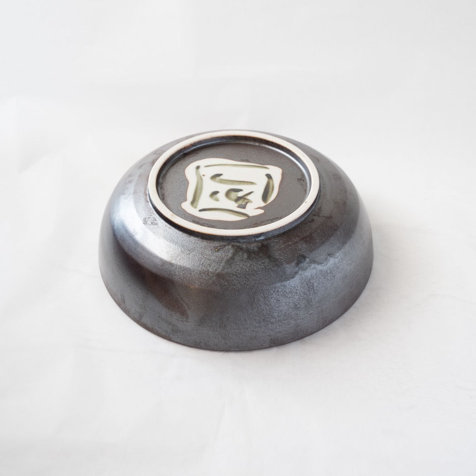 Underside view of a Shigaraki iron-glazed ceramic bowl by Katsuhiko Sato, showcasing a circular foot ring and a white slip square painted with a dark abstract character.