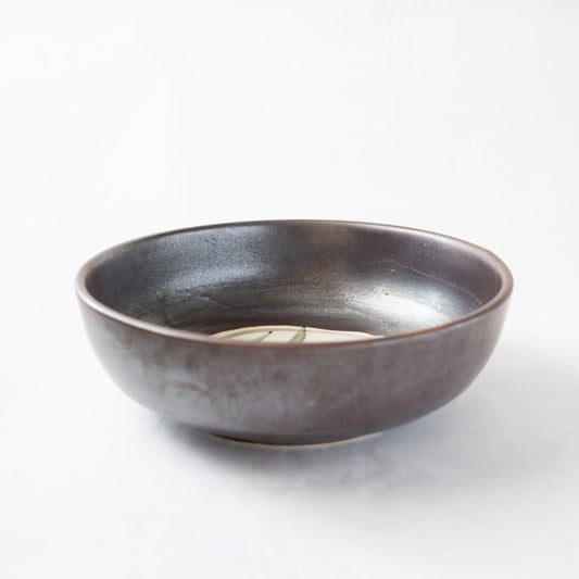 A detailed angled top-view of the Shigaraki iron-glazed confectionery bowl by Katsuhiko Sato, showcasing its dark, metallic-sheen profile and a glimpse of the internal white slip patch with its minimalist linear drawing.