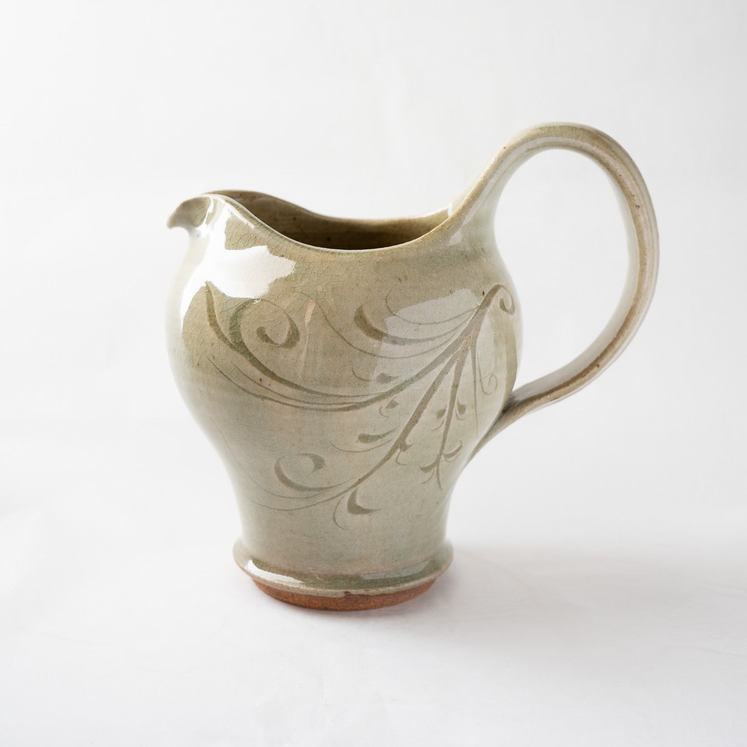 Profile view of the ash-green ceramic pitcher against a soft white background, capturing its full sculptural silhouette, light reflections on the glossy glaze, and detailed line-work.