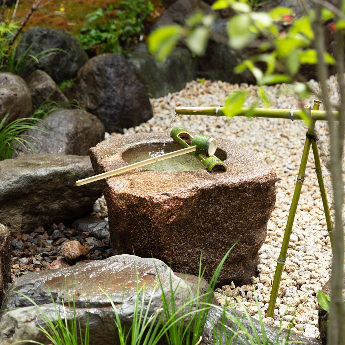 Low stone water basin (tsukubai) with a bamboo ladle, set among moss, gravel, and natural stones in a Japanese tea garden.