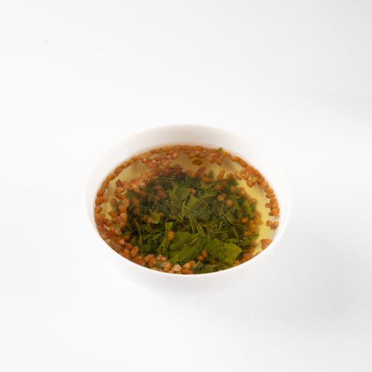 Brewed pesticide-free Genmaicha tea in a white porcelain bowl, showing unfolded green tea leaves and floating golden roasted rice, delivering a cozy warmth similar to roasted Hojicha.