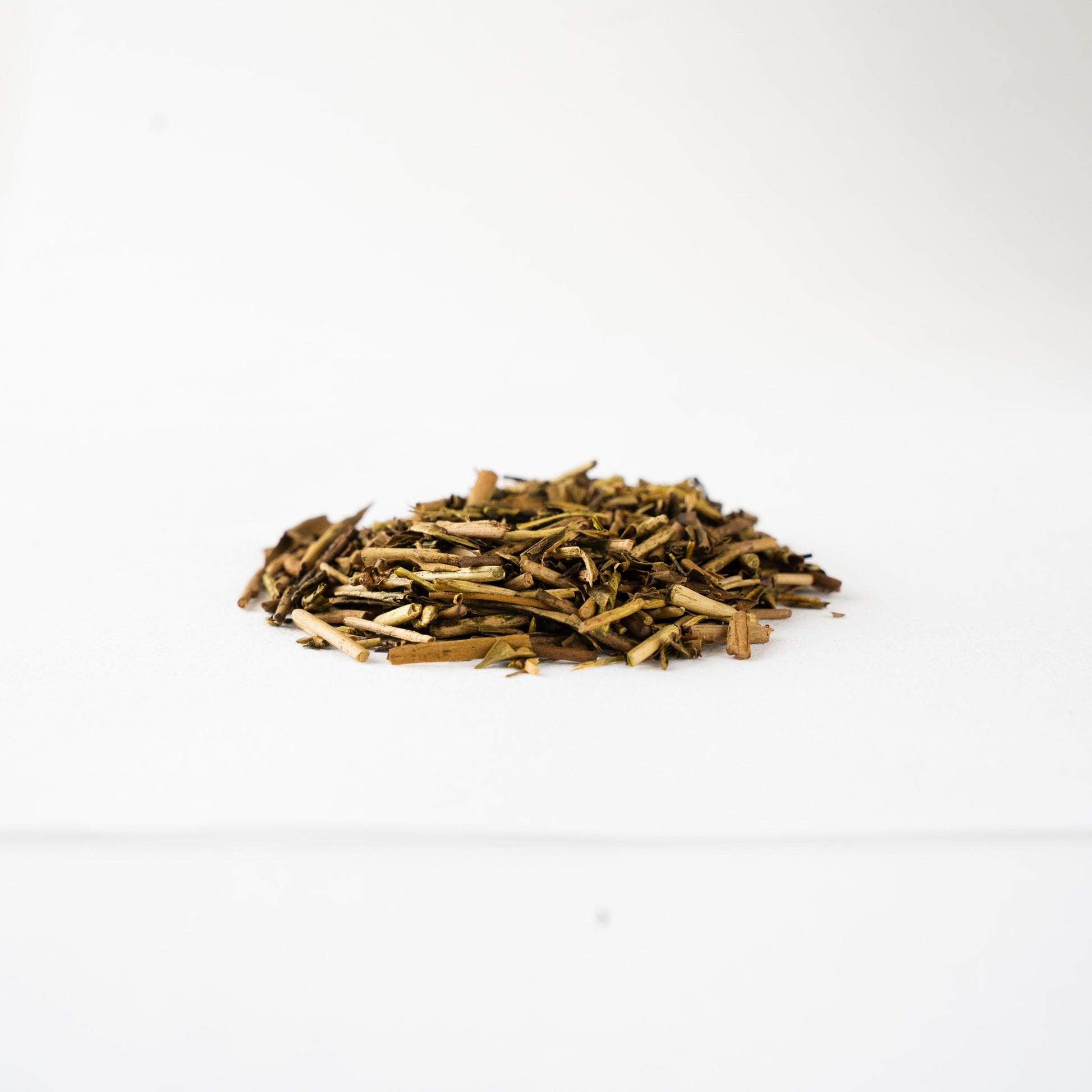 Pile of roasted first-flush hojicha tea leaves showing earthy brown tones and delicate twig textures against a white background.