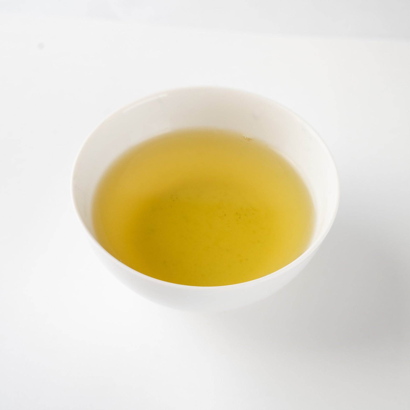 A serene top-down perspective of a clear yellowish-green infusion of Japanese green tea, held within a simple, minimalist white ceramic bowl, revealing fine tea particles.