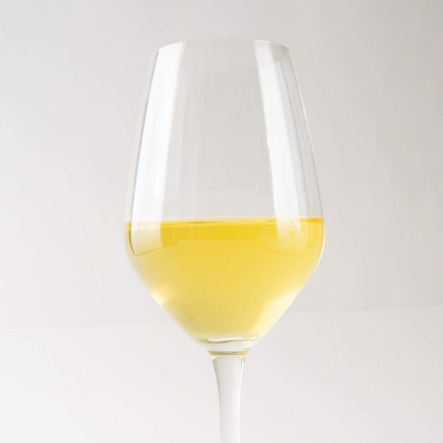 A serene front-on portrait view of an elegant, slender-stemmed, crystal-clear wine glass, set against a simple, minimalist white background. The glass is filled halfway with a clear, vibrant, yellowish-gold infusion of white wine. The natural light highlights the transparent glass and illuminates the vivid hue and clarity of the liquid, creating a sense of refined elegance and sophisticated enjoyment.