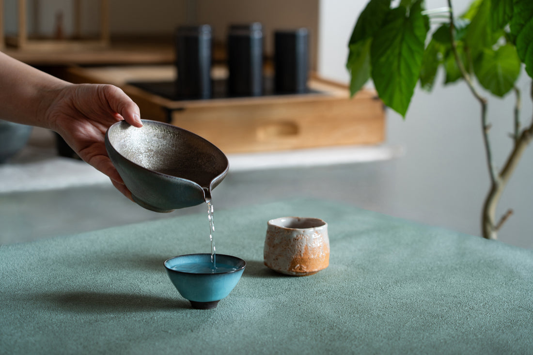 An elegant moment of focus and flow. An elegant hand guides water from a spouted katakuchi into a turquoise guinomi, set upon a tactile green surface within a refined, contemporary interior.