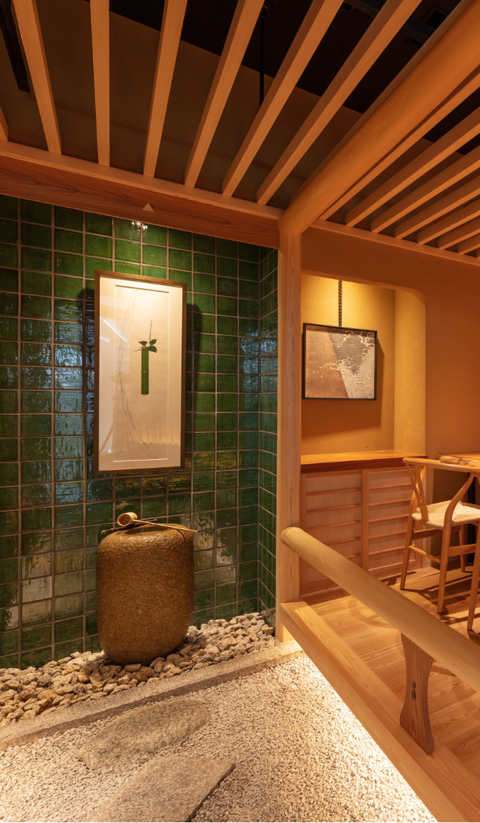 Soft ambient light illuminates an interior courtyard featuring a stone basin (tsukubai) set against deep green glazed tiles, framed by exposed timber rafters and warm earth-plastered walls.