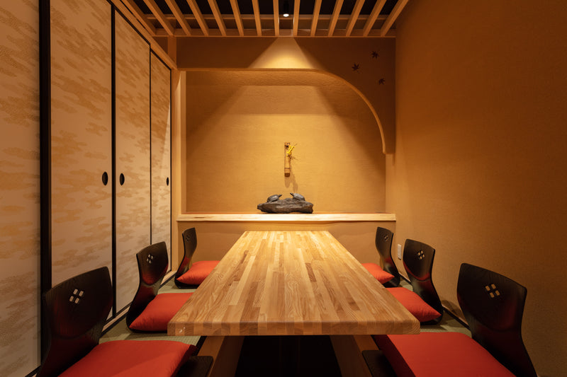 A sunken horigotatsu room framed by gold-accented sliding fusuma screens, an earthen plaster alcove, and a long natural timber table illuminated by soft, directional spotlighting.