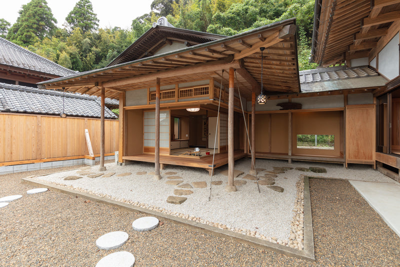 A tranquil tea pavilion nestled against lush greenery, where stepping stones across white gravel lead into a softly lit tatami room framed by natural wood.