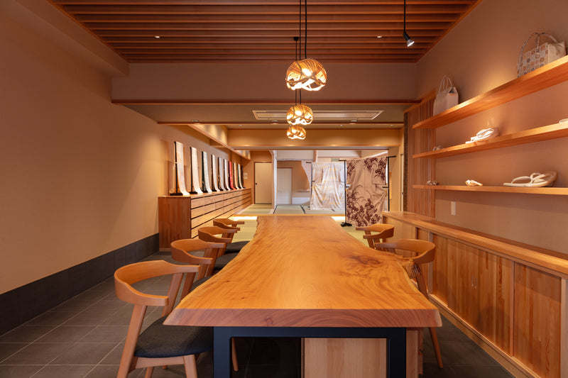 A commanding live-edge timber table anchors an elegant textile showroom, where warm ambient light highlights traditional kimono craftsmanship and natural wood joinery.