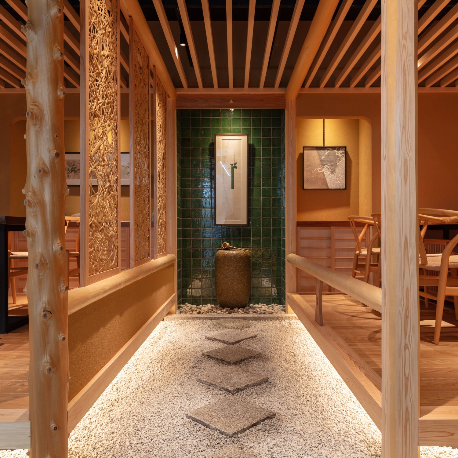 A modern Japanese interior path featuring a white gravel walkway with diamond-shaped stepping stones, natural timber pillars, woven screens, and a deep green tiled focal wall with a stone water basin and hanging scroll.