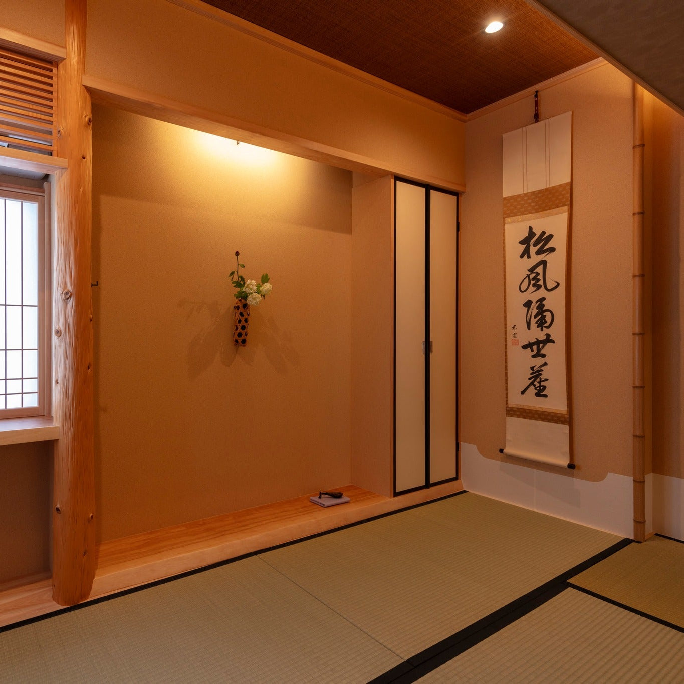 A softly lit traditional Japanese tearoom featuring a wide tokonoma alcove with a wall-mounted bamboo flower vase, a calligraphy hanging scroll on the adjacent wall, and tatami mat flooring.