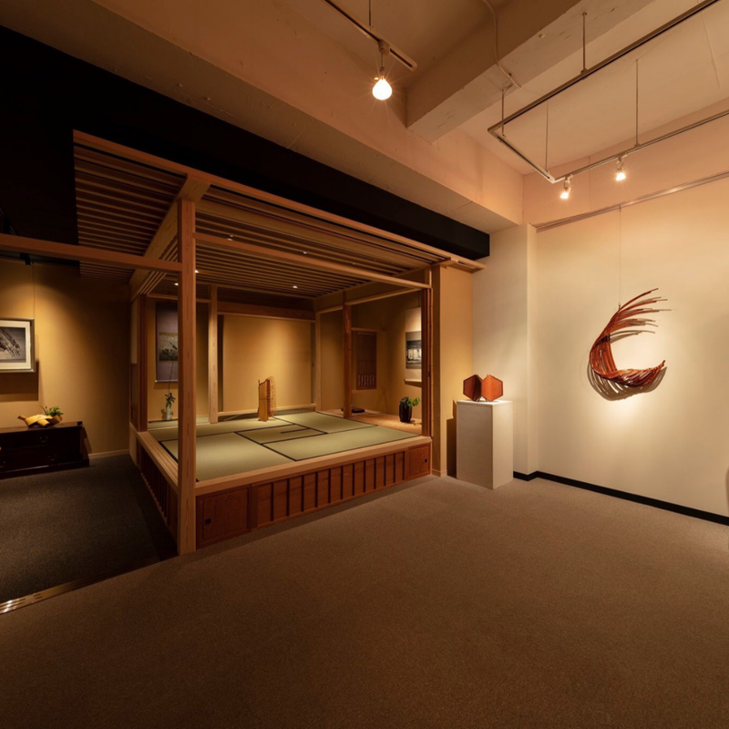 An open-framed timber tea pavilion with tatami flooring sits within a modern gallery space, anchored by contemporary bamboo sculpture and warm, focused spotlighting.