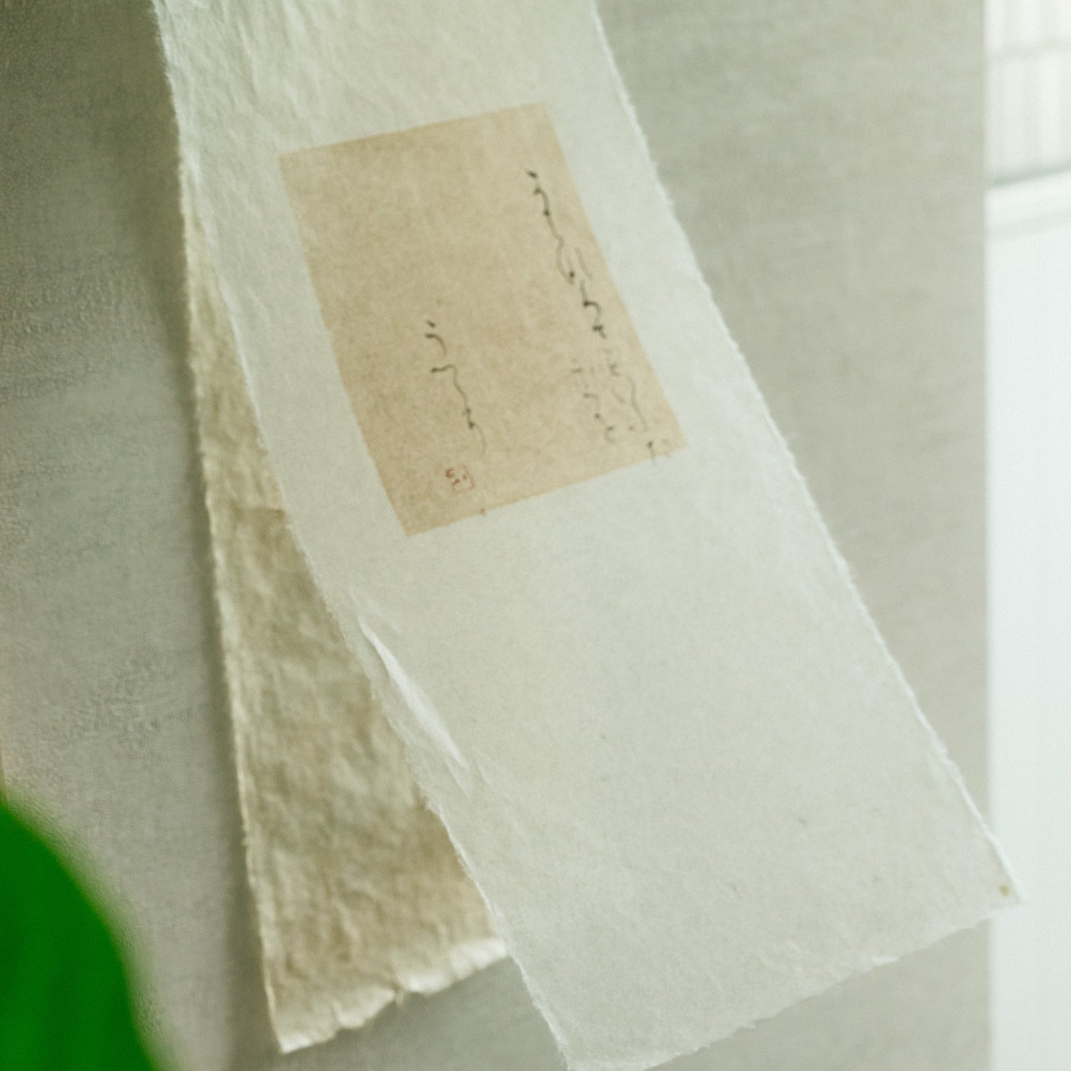 Hand-molded washi paper with soft deckled edges, bearing a fragment of delicate Japanese calligraphy and a red seal mark against a quiet, textured backdrop.