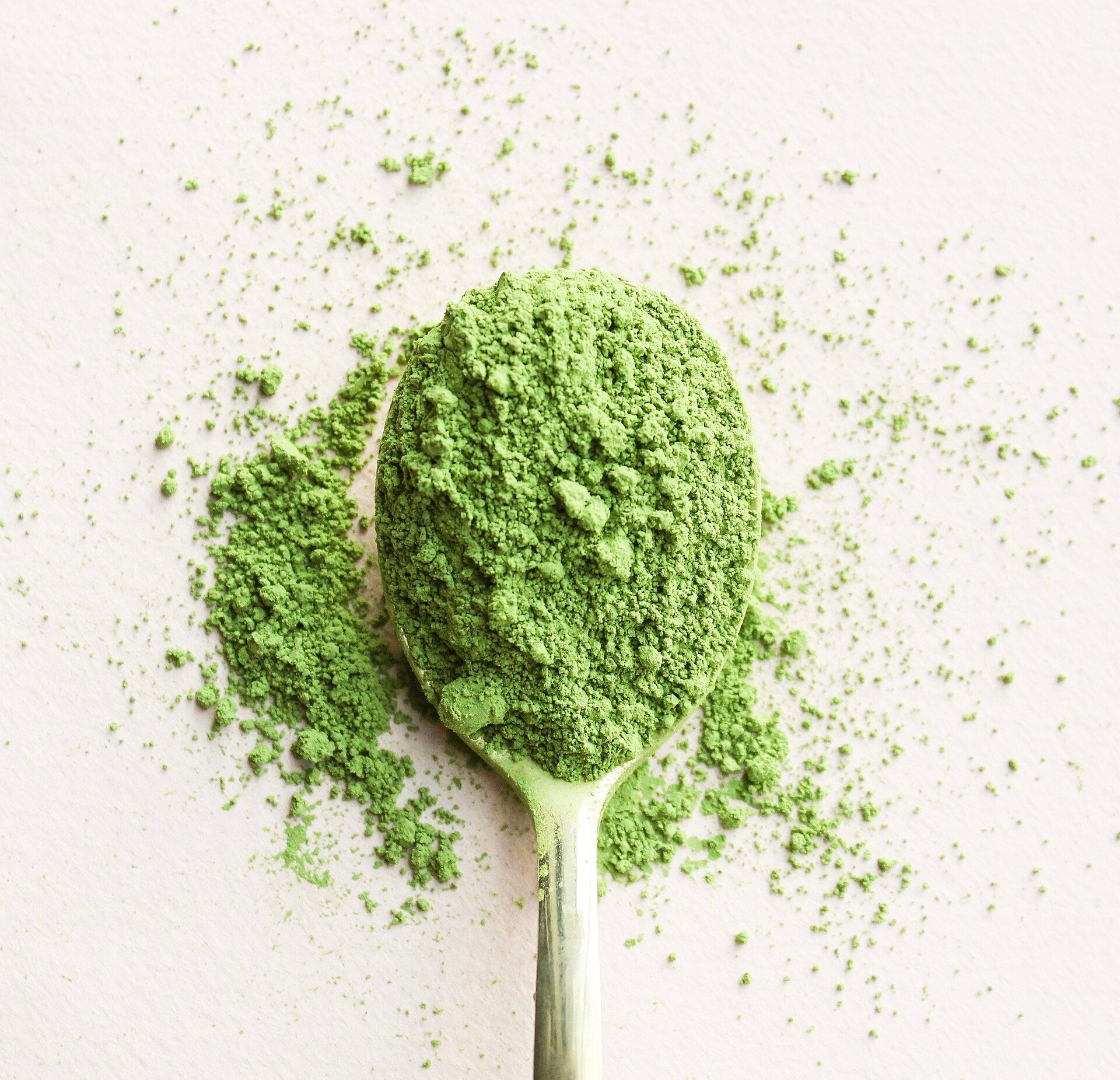 A spoon delicately filled with vibrant green matcha powder, surrounded by a fine dusting of tea particles on a textured, light-colored background.
