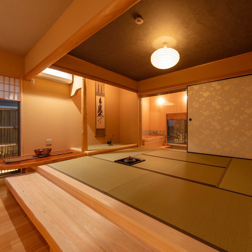 A serene and refined modern Japanese-style tatami bedroom, featuring a central futon, a tokonoma alcove with a hanging scroll, decorative sliding doors, and a glimpse of an integrated wooden bath in the background.