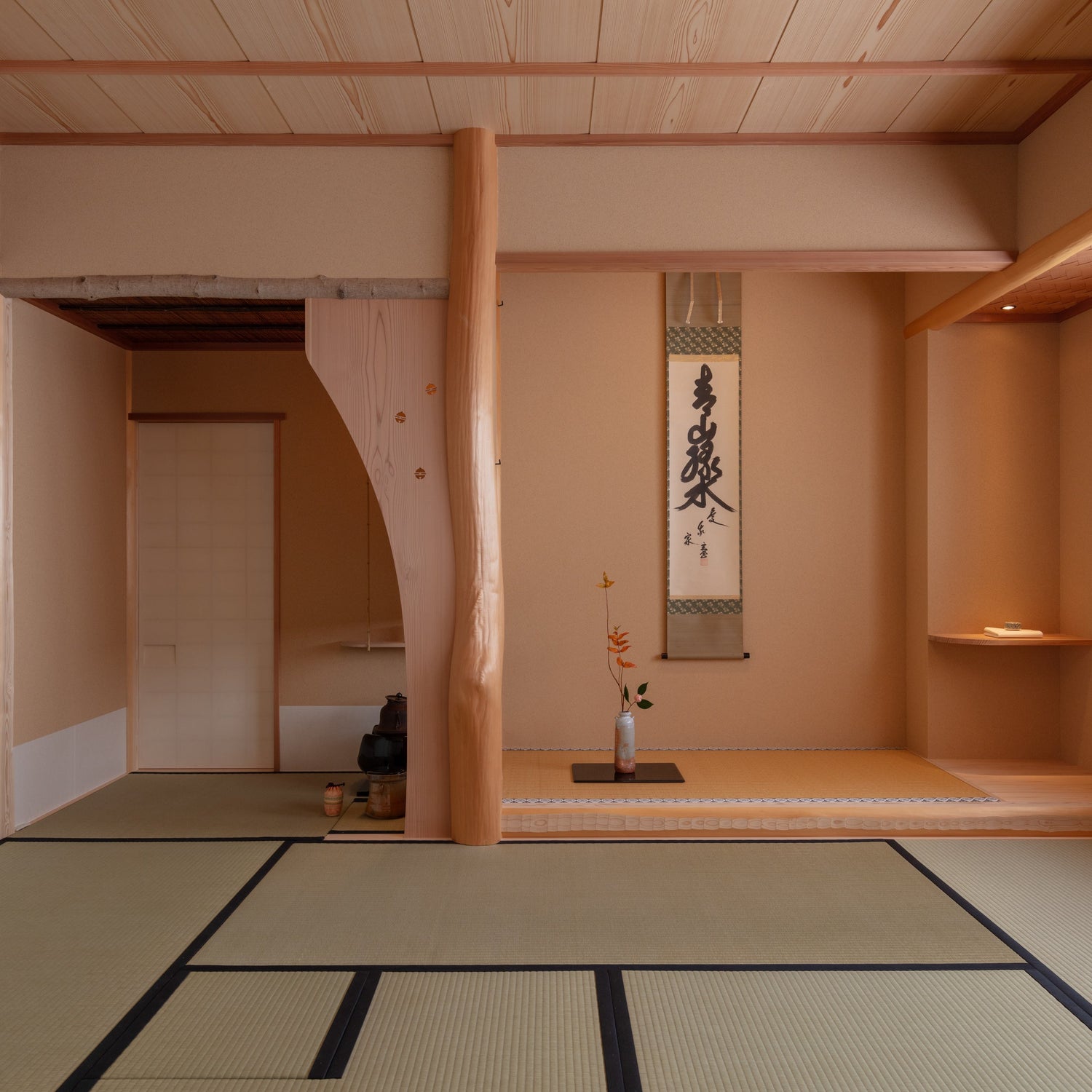 A traditional Japanese tea room (chashitsu) featuring a raised tokonoma alcove with a hanging calligraphy scroll, a seasonal flower arrangement, a natural log pillar, and tatami mat flooring.