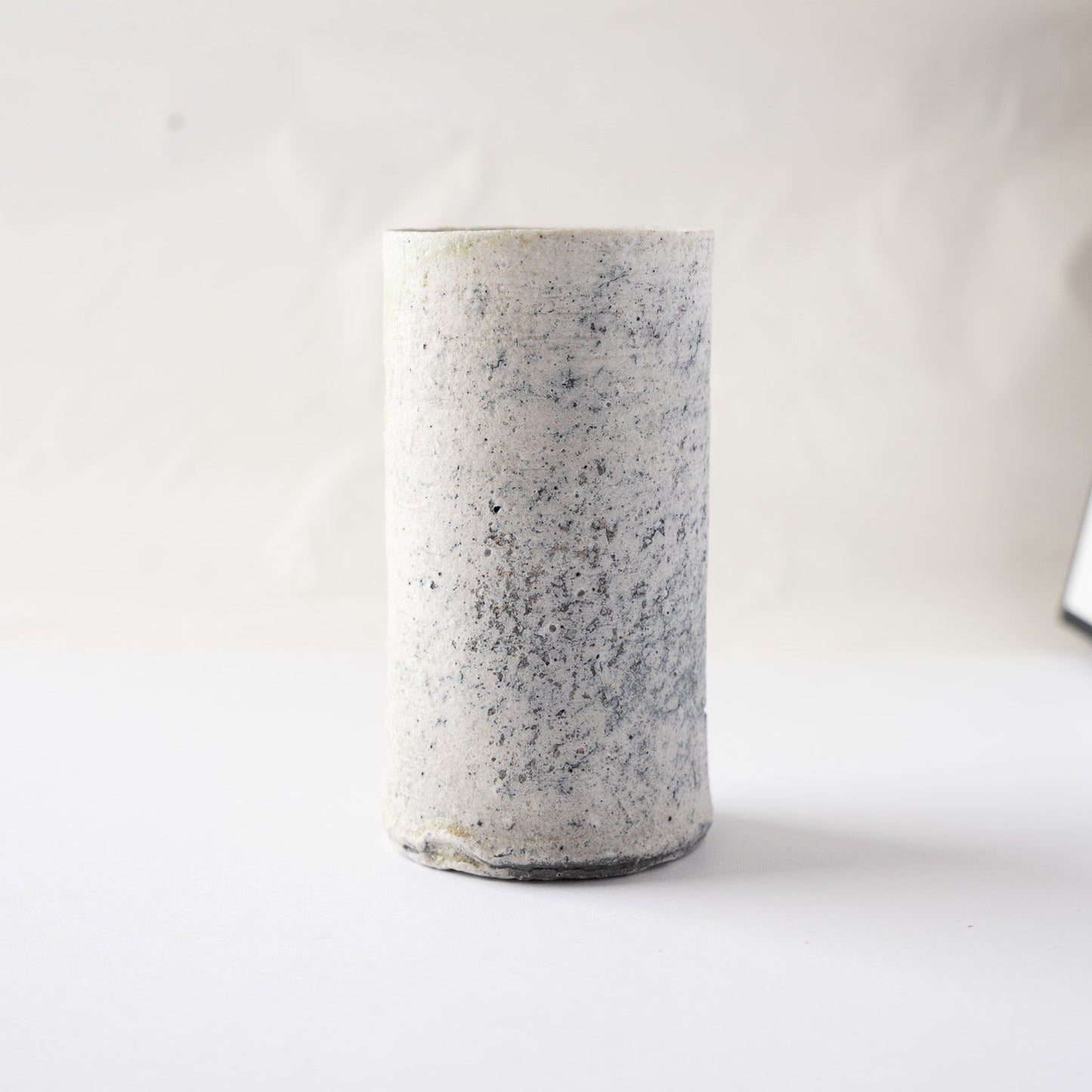 Full view of the pristine white side of the cylindrical YUNOMI cup by Yui Tsujimura, showing its column-like architectural form and organic spotting.