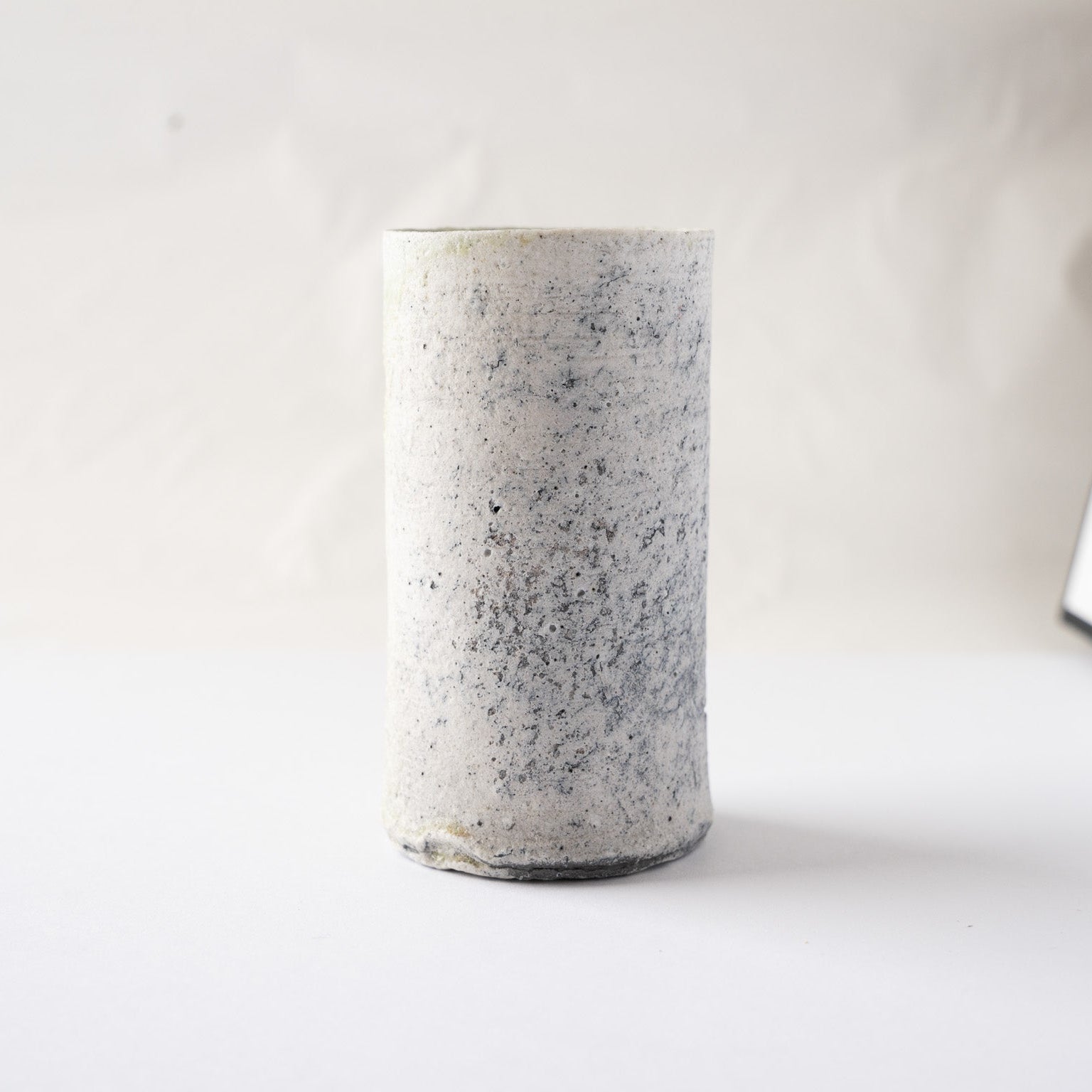 Full view of the pristine white side of the cylindrical YUNOMI cup by Yui Tsujimura, showing its column-like architectural form and organic spotting.