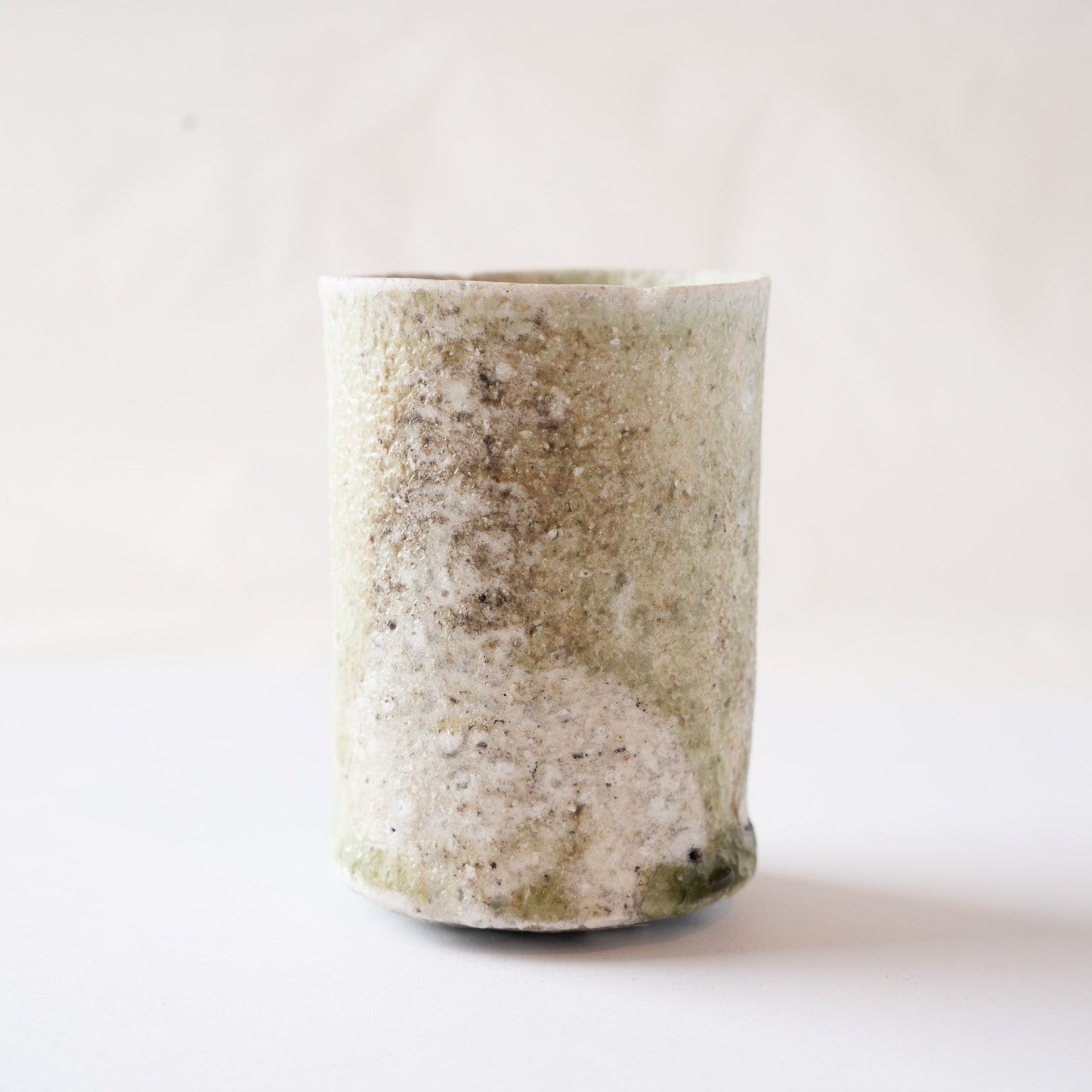 Front view of a cylindrical natural glaze YUNOMI cup by Yui Tsujimura, featuring a textured beige and tan surface with a prominent olive-green vidro droplet at the bottom right.