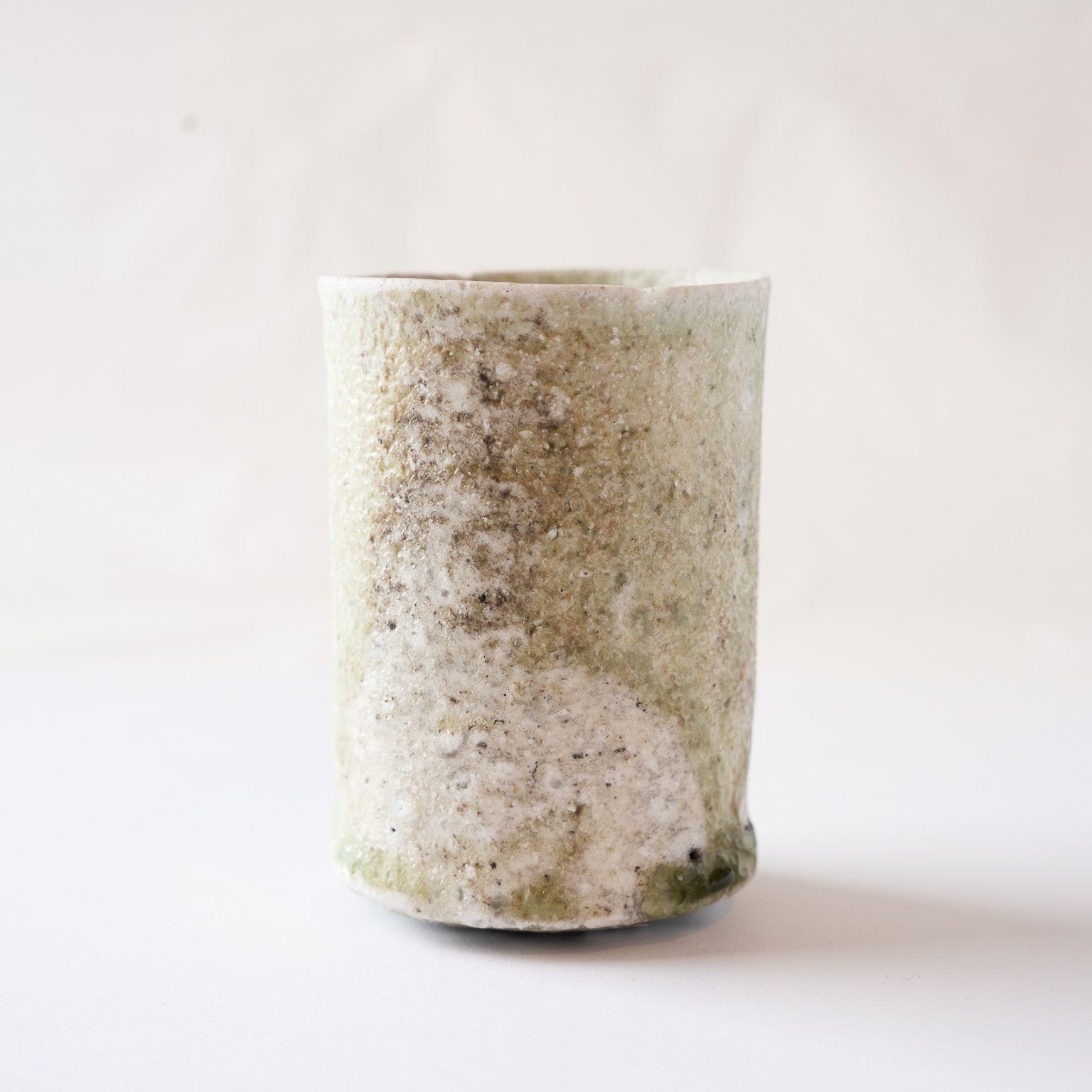 Front view of a cylindrical natural glaze YUNOMI cup by Yui Tsujimura, featuring a textured beige and tan surface with a prominent olive-green vidro droplet at the bottom right.