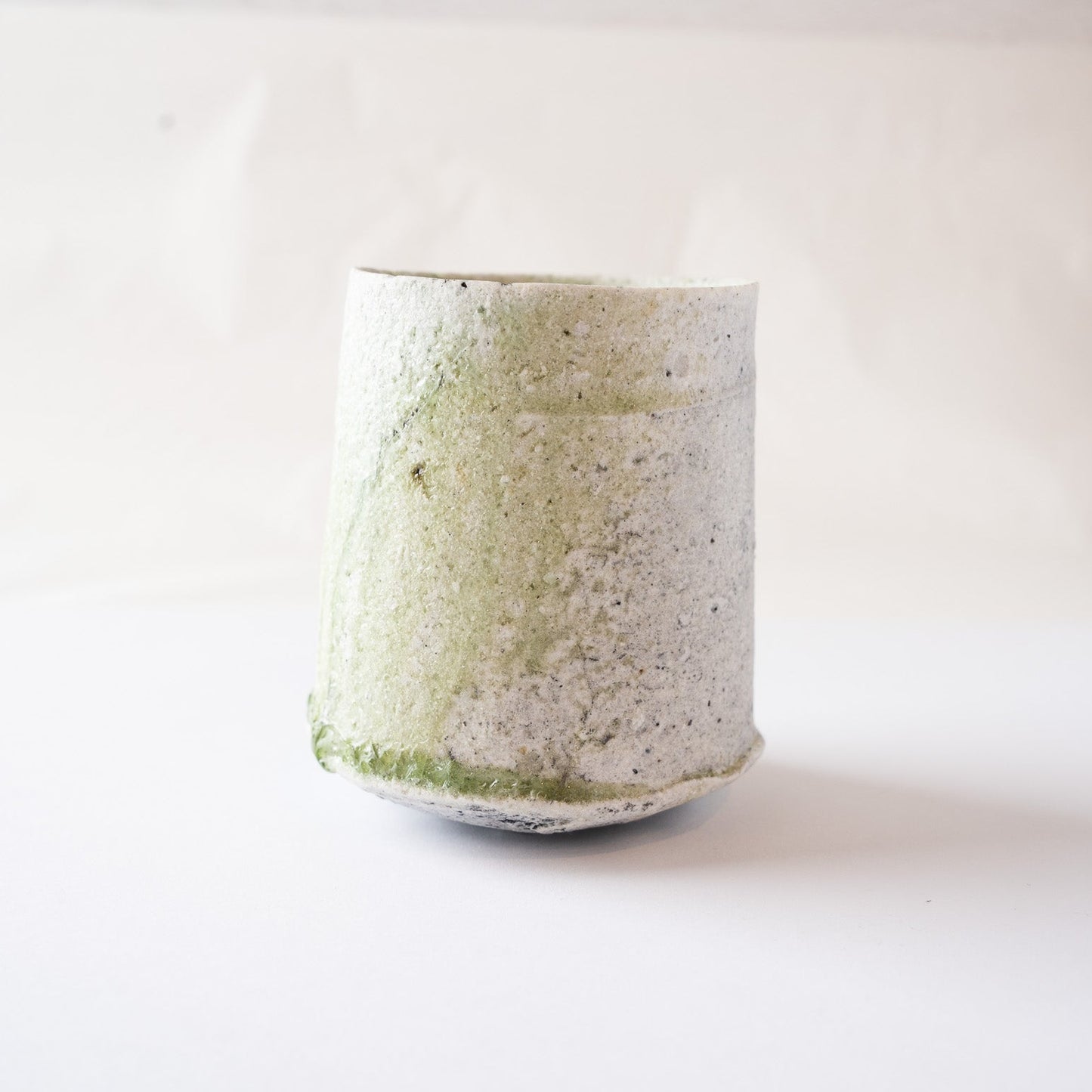 Profile view of a cylindrical natural glaze YUNOMI cup by Yui Tsujimura, showcasing rough, matte white crystalline ash deposits on the upper body, ceiled at the base by a prominent, vitrified ring of deep emerald green vidro glass.