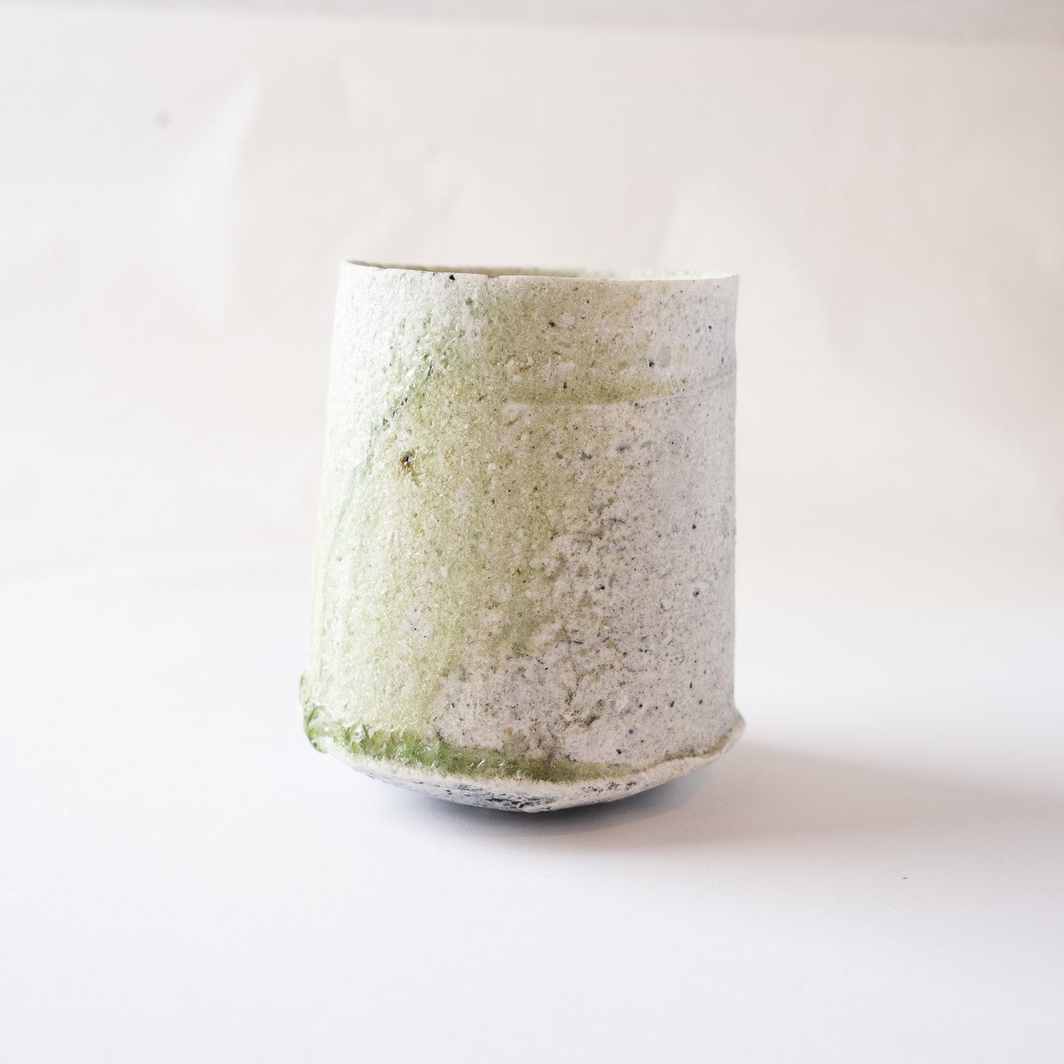 Profile view of a cylindrical natural glaze YUNOMI cup by Yui Tsujimura, showcasing rough, matte white crystalline ash deposits on the upper body, ceiled at the base by a prominent, vitrified ring of deep emerald green vidro glass.